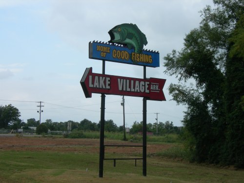 Sign for Lake Village, Arkansas, featuring a fish graphic and arrows pointing to "Good Fishing."