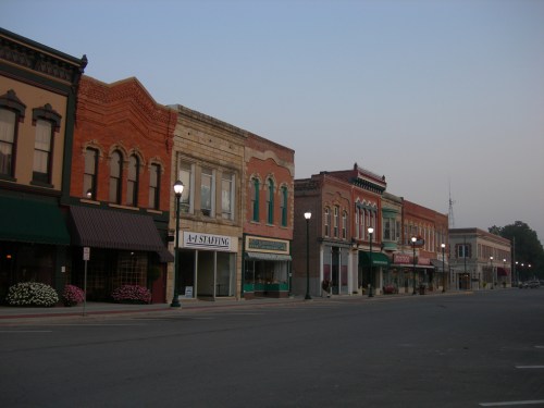A quiet street lined with historic brick buildings and storefronts at dusk, with streetlights illuminating the scene.