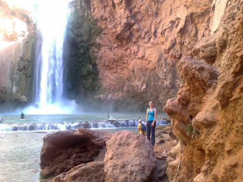 A person stands near a waterfall surrounded by rocky terrain and lush greenery, with water cascading in the background.