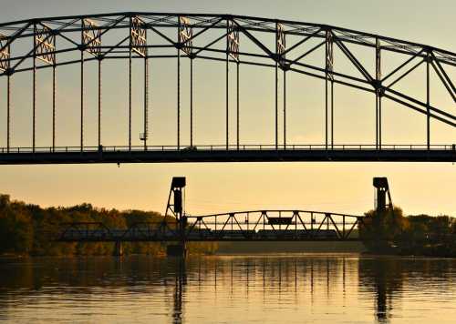 Two bridges span a calm river at sunset, with reflections shimmering on the water's surface.