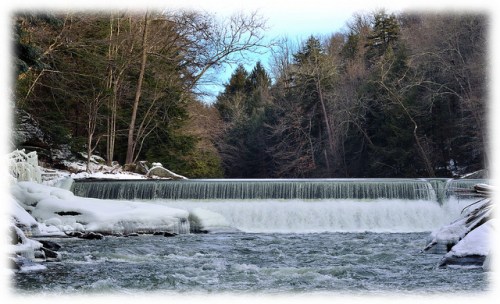 A serene winter scene of a waterfall cascading over a frozen ledge, surrounded by trees and icy banks.
