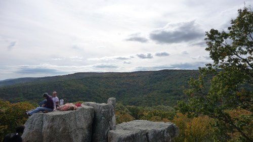 Two people sit on a large rock overlooking a lush, green valley under a cloudy sky.
