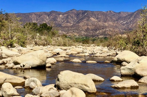 A serene river flows through a rocky landscape, with mountains in the background under a clear blue sky.