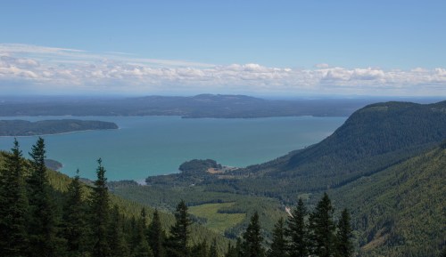 A panoramic view of a serene lake surrounded by lush green forests and distant mountains under a clear blue sky.