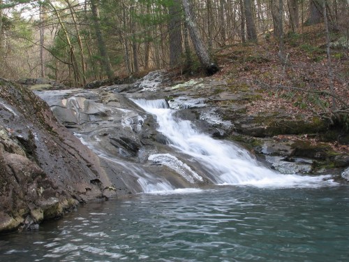 A serene waterfall cascades over rocky terrain, surrounded by lush trees and a calm pool of water below.