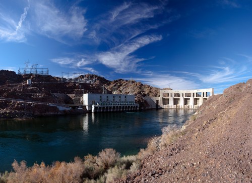 A panoramic view of a dam along a river, surrounded by rocky terrain and a clear blue sky with wispy clouds.
