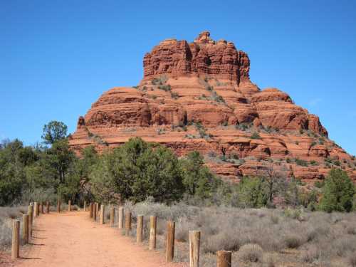 A dirt path lined with wooden posts leads to a red rock formation under a clear blue sky.