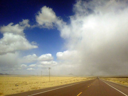 A wide, empty road stretches through a desert landscape under a cloudy sky, with distant mountains on the horizon.