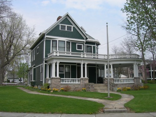 A large green Victorian-style house with a wraparound porch and landscaped yard on a cloudy day.