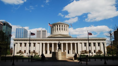 Ohio Statehouse with a dome, surrounded by flags and trees, under a blue sky with clouds.