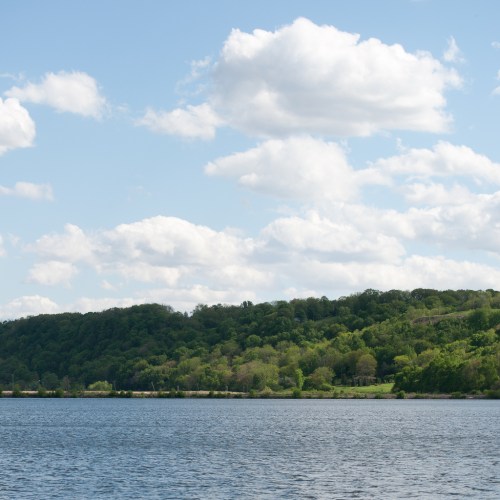A serene landscape featuring a calm lake, lush green hills, and a blue sky with fluffy white clouds.