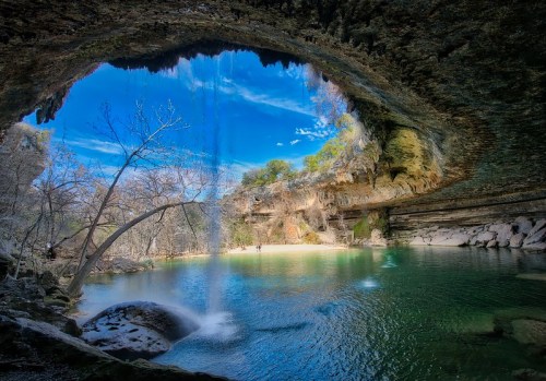 A serene cave with a waterfall cascading into a clear pool, surrounded by trees and rocky formations under a blue sky.