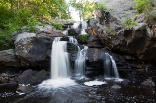 A serene waterfall cascading over rocks into a dark pool, surrounded by lush greenery and natural stone formations.
