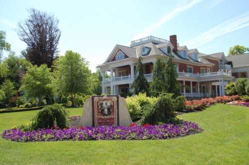 A large, elegant house surrounded by lush greenery and colorful flower beds on a sunny day.