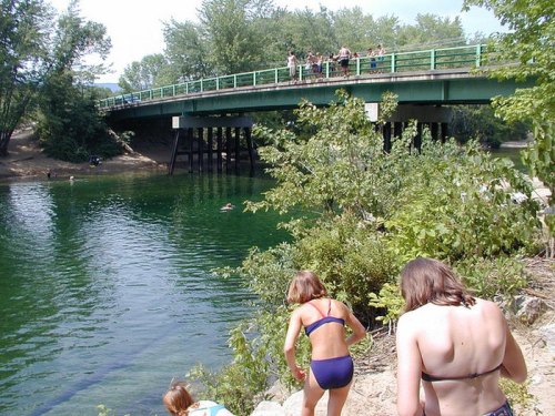 A group of people near a green bridge, with some swimming in a clear river and others climbing the bank.