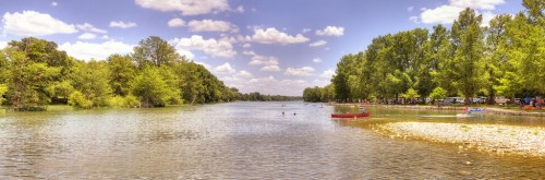 A serene river scene with lush trees, people swimming, and kayaks under a bright blue sky with fluffy clouds.