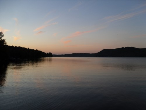 A serene lake at sunset, with calm waters reflecting soft pink and orange hues in the sky and surrounding hills.