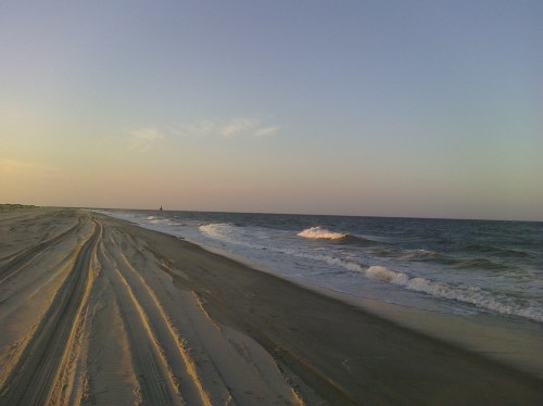 A serene beach scene at sunset, with gentle waves and tire tracks in the sand leading towards the horizon.
