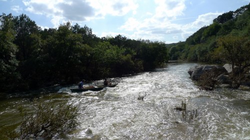 A serene river scene with two people in a boat surrounded by lush greenery and a bright, cloudy sky.