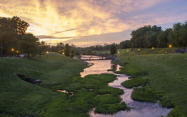 A serene river flows through a green landscape at sunset, with trees and soft lighting along the banks.