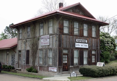 A rustic two-story building with wooden siding, signs for "Raymond" and "Antiques," surrounded by greenery.