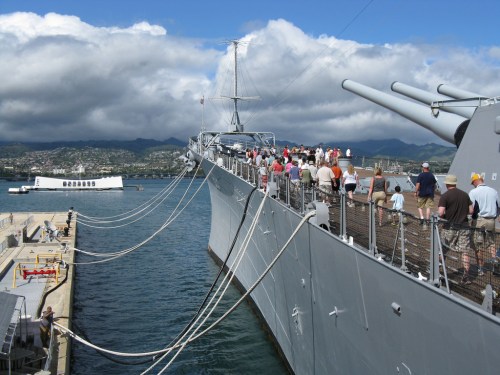 A battleship docked at a harbor with visitors walking along its deck under a cloudy sky.