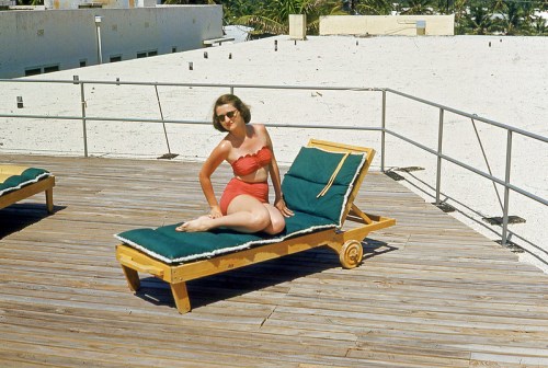 A woman in a red swimsuit relaxes on a lounge chair on a rooftop deck, enjoying the sunny day.