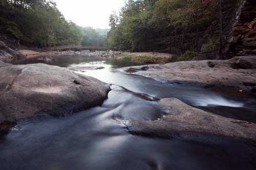 A serene river flows over smooth rocks, surrounded by lush trees and a distant wooden bridge in a tranquil landscape.