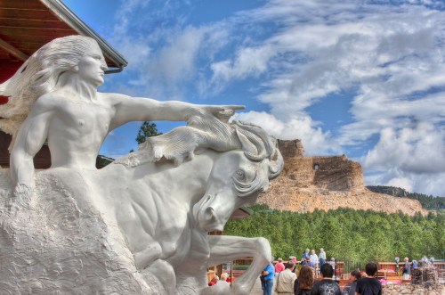 A large white statue of a man on a horse, pointing forward, with a scenic backdrop of cliffs and a cloudy sky.
