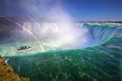 A boat near the edge of Niagara Falls, with mist and vibrant rainbows in the background.