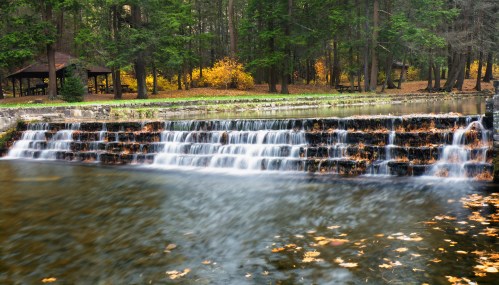 A serene waterfall cascades over rocks into a calm pond, surrounded by lush trees and autumn foliage.