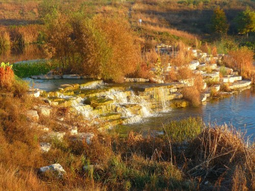A serene landscape featuring a small waterfall cascading over rocks into a calm pond, surrounded by autumn foliage.