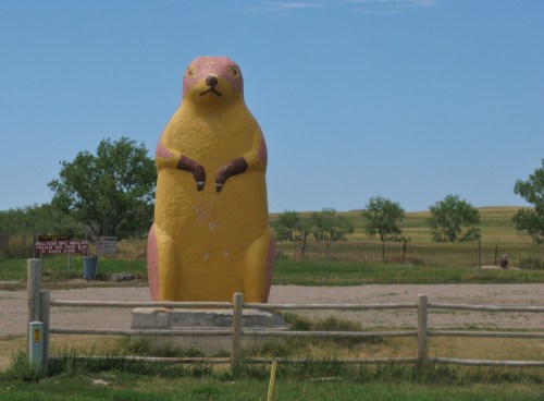 A large, colorful statue of a prairie dog stands in a grassy area with trees and blue sky in the background.