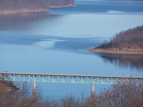 A serene view of a blue bridge spanning a calm lake, surrounded by wooded hills and clear skies.