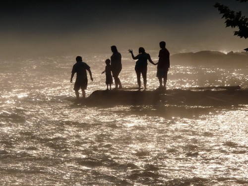 Silhouettes of a family standing on a rocky shore, with shimmering water and mist in the background.