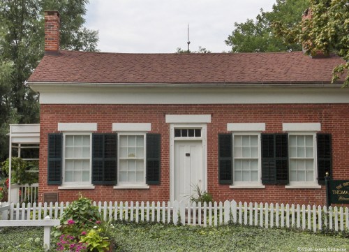 A brick house with white trim, black shutters, and a picket fence, surrounded by greenery and flowers.