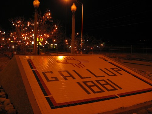A nighttime view of a tiled sign reading "GALLUP 1881," surrounded by decorative lights and a tree.