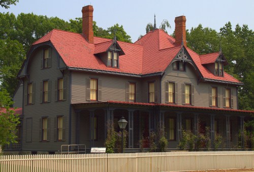 A large, historic gray house with a red roof, featuring multiple chimneys and a white picket fence.