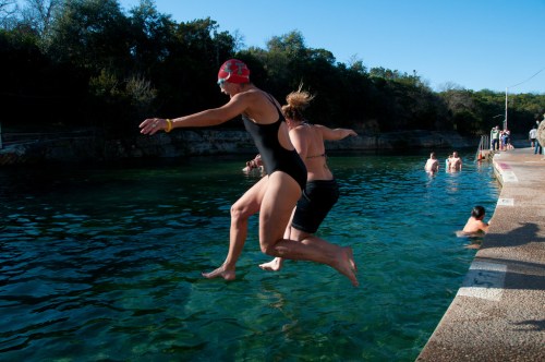 Two people jump into a clear blue swimming area, with others swimming nearby on a sunny day.