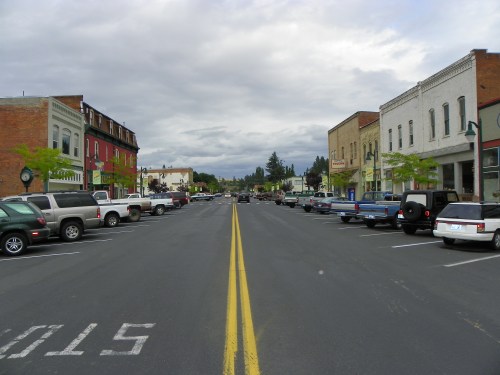 A quiet downtown street lined with parked cars and historic buildings under a cloudy sky.