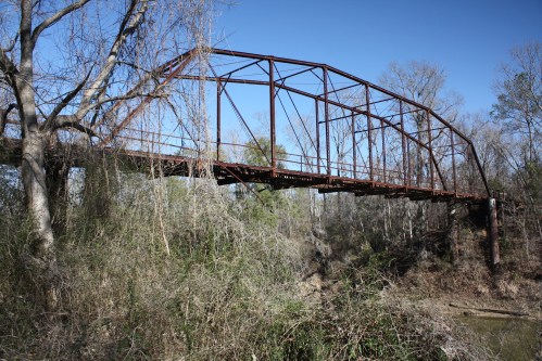 A rusted metal bridge spans a creek, surrounded by trees and overgrown vegetation under a clear blue sky.