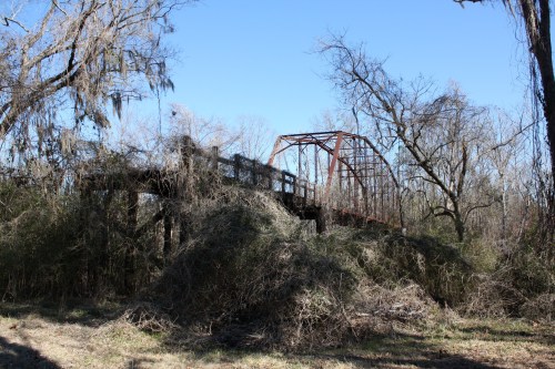 An overgrown, abandoned bridge surrounded by trees and dense vegetation under a clear blue sky.