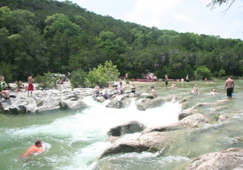 People swimming and relaxing in a natural river setting with rocky banks and lush greenery in the background.
