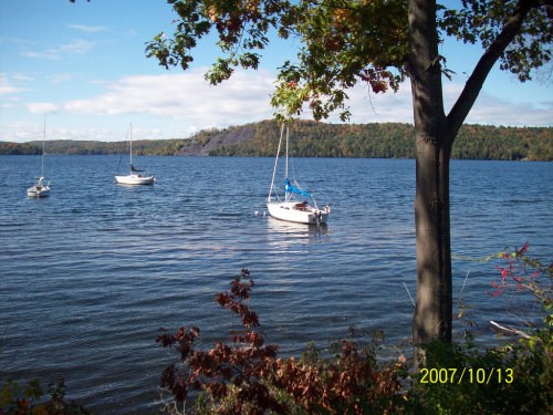 A serene lake scene with sailboats on the water, surrounded by trees and autumn foliage under a clear blue sky.