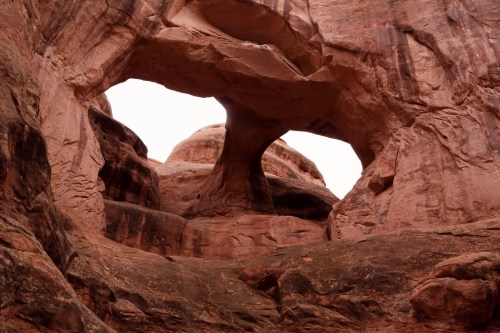 Two natural rock arches framed by rugged red rock formations under a cloudy sky.