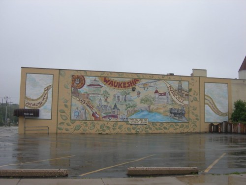 Mural on a building in Waukesha, featuring local landmarks and scenic elements, under a gray, overcast sky.