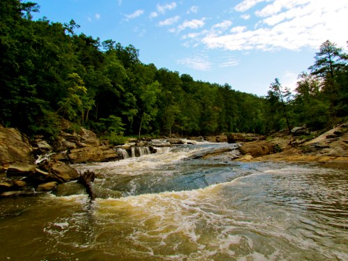 A serene river flows over rocky terrain, surrounded by lush green trees under a blue sky with scattered clouds.