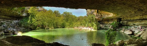 A serene natural pool surrounded by lush greenery, viewed from a rocky overhang. People enjoy the water below.