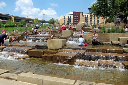 People enjoying a water feature with cascading waterfalls, surrounded by greenery and a hotel in the background.