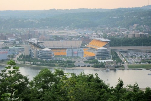 Aerial view of a stadium by a river, surrounded by green hills and city buildings in the background.